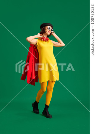 Portrait of pretty woman wearing fashion look with sunglasses and headphones holds red jacket isolated bright green studio background. 108780188