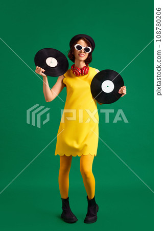 Young pretty woman wearing vivid fashion look with sunglasses and hat holds retro music plates isolated bright green studio background. 108780206
