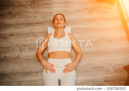 Adult athletic woman, in white bodysuit, performing fascia exercises on the floor - caucasian woman using a massage foam roller - a tool to relieve tension in the back and relieve muscle pain - the Adult athletic woman, in white bodysuit, performing fascia exercises on the floor - caucasian woman using a massage foam roller - a tool to relieve tension in the back and relieve muscle pain - the 108780565
