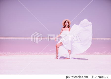 Woman in pink salt lake. She in a white dress and hat enjoys the scenic view of a pink salt lake as she walks along the white, salty shore, creating a lasting memory. 108780627