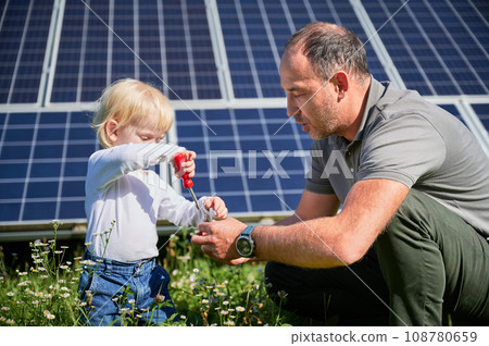 Dad showing to his little son how to use screwdriver in background of solar panels. Father teaching his child how to use tools. Son and his young dad learning tools. 108780659