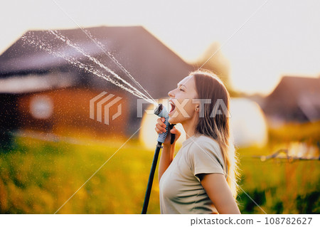 Watering vegetables from garden hose, young woman splashes water while fooling around and making faces. Watering vegetables from garden hose, young woman splashes water while fooling around and making faces. 108782627