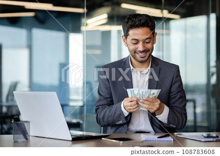 Happy Arab worker in office sitting in front of laptop contentedly counting dollars earned online, success concept, big lottery win. Happy Arab worker in office sitting in front of laptop contentedly counting dollars earned online, success concept, big lottery win. 108783608