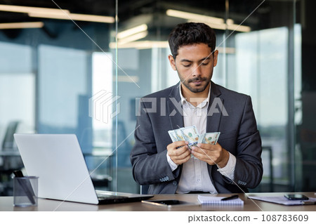 Serious Arab businessman sits in a modern office at a table in front of a laptop, counts earned money, online earnings, a wad of dollars in his hands. Serious Arab businessman sits in a modern office at a table in front of a laptop, counts earned money, online earnings, a wad of dollars in his hands. 108783609
