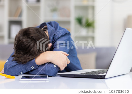 Tired student boy sitting at desk in front of laptop during distance lesson, exhausted sleepy teenager lying on hands on desk with head down, tired of studying. 108783650