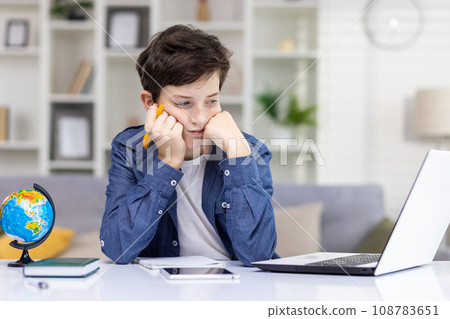 Boy teenager who is bored, sitting at the laptop, studying remotely at home, holding his head with his hands, tired from studying, listening to the lesson. 108783651