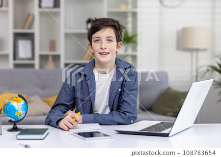Portrait of happy teenage boy student sitting at table at home, studying via distance learning, doing homework, smiling, looking at camera. 108783654
