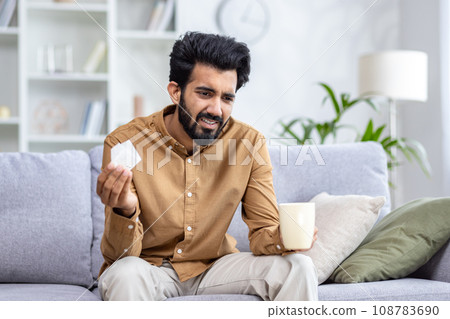 Sick Hindu man sitting on sofa at home, holding cup in hand, drinking tea, holding napkin, viral diseases, runny nose and cough, health concept. Sick Hindu man sitting on sofa at home, holding cup in hand, drinking tea, holding napkin, viral diseases, runny nose and cough, health concept. 108783690
