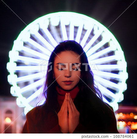 young woman with closed eyes puts her hands together in front of a ferris wheel 108784061