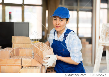 Female builder carrying hollow clay bricks and putting them on brick stack 108785574
