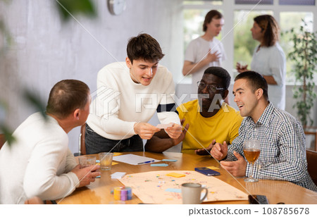 Excited guy playing board game at table with male friends Excited guy playing board game at table with male friends 108785678