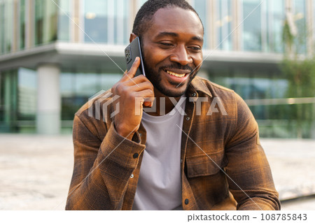 African american man talking on smartphone on street in city outdoor. Man with cell phone chatting with friends. Smiling person making answering call by cellphone. Guy having conversation by mobile African american man talking on smartphone on street in city outdoor. Man with cell phone chatting with friends. Smiling person making answering call by cellphone. Guy having conversation by mobile 108785843