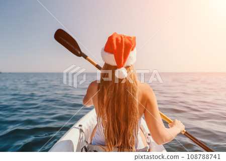 Woman in kayak back view. Happy young woman in Santa hat floating in kayak on calm sea. Summer holiday vacation and cheerful female people relaxing having fun on the boat. 108788741