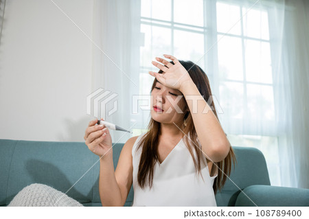 Sick Asian young woman sit under blanket on sofa she sneeze checking temp with digital thermometer and tissue paper in living room at home, Cold flu and migraine concept, medical healthcare 108789400