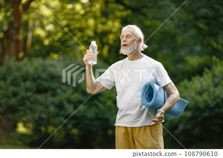 Senior man with yoga mat standing at park with bottle of water. Positive mature man after fitness or yoga class in nature. Wellness and healthy lifestyle on retirement. 108790610