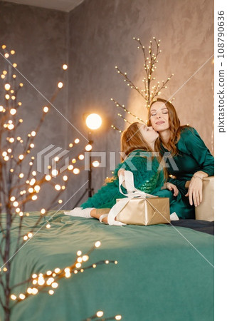 Portrait of a little girl celebrating Christmas and opening a gift box. Girl and her mother posing for a photo. Kid and woman wearing green dresses. 108790636