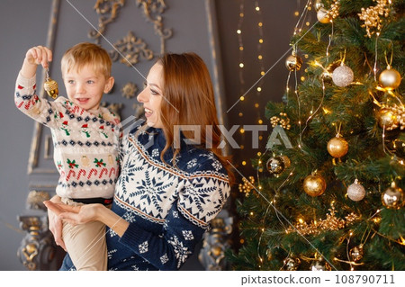 Portrait of a mother holding her son on hands. Young mother with little boy in studio posing for a photo. Woman and kid wearing knitted sweaters with Christmas prints. 108790711