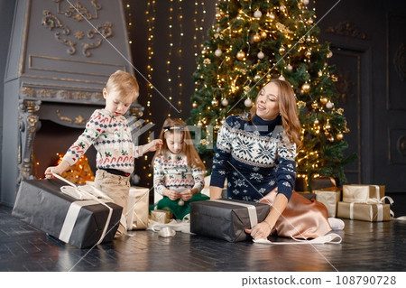 Young mother, little daughter and son in studio posing for a photo. Woman and kids wearing knitted sweaters with Christmas prints. Family celebrating Christmas together. 108790728