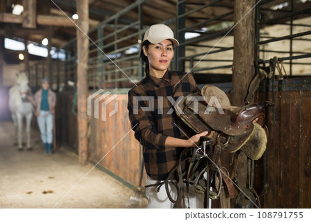 Woman horse breeder carrying leather saddle 108791755
