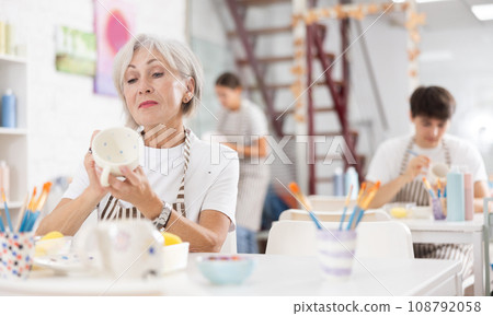 Talented senior female artisan applying design to ceramic mug in pottery studio 108792058
