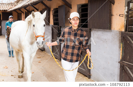 Positive woman jockey leads white horse by the bridle on the street along stable 108792113