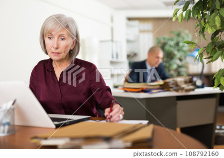 Concentrated elderly female office employee working with laptop Concentrated elderly female office employee working with laptop 108792160