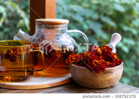 Marigold tea still life on table in green garden background. Healthy hot drink benefits. Natural organic aromatic drink in cup. Home-grown immunity-boosting herbs for tea. Autumn winter warming drink Marigold tea still life on table in green garden background. Healthy hot drink benefits. Natural organic aromatic drink in cup. Home-grown immunity-boosting herbs for tea. Autumn winter warming drink 108792378