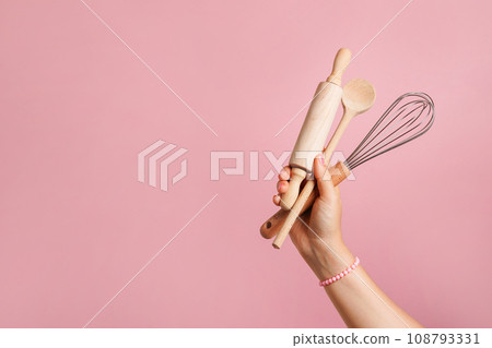 Female hand holding kitchen utensils for food and bakery on pink background. whisk, rolling pin and wooden spoon 108793331