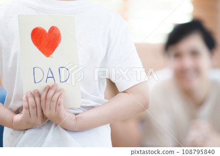 Asian lovely little boy happy to give his dad a Father's Day greeting card and present while living together in living room, diverse gender and lifestyle concept. Asian lovely little boy happy to give his dad a Father's Day greeting card and present while living together in living room, diverse gender and lifestyle concept. 108795840