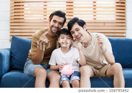Happy LGBTQ+ gay family with adopted little son stay together in living room and teaching their adopted son about wealth and saving money for future. Diversity in gender and lifestyle concept. Happy LGBTQ+ gay family with adopted little son stay together in living room and teaching their adopted son about wealth and saving money for future. Diversity in gender and lifestyle concept. 108795842
