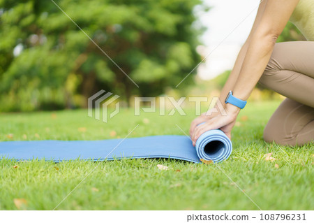 Happy beautiful Asian woman rolling out an exercise - yoga mat on the grass field to prepare doing a yoga and mediation exercise. Woman preparing a yoga or body weight and meditating exercise. 108796231
