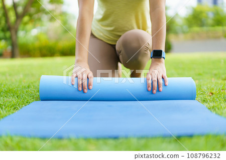 Happy beautiful Asian woman rolling out an exercise - yoga mat on the grass field to prepare doing a yoga and mediation exercise. Woman preparing a yoga or body weight and meditating exercise. Happy beautiful Asian woman rolling out an exercise - yoga mat on the grass field to prepare doing a yoga and mediation exercise. Woman preparing a yoga or body weight and meditating exercise. 108796232