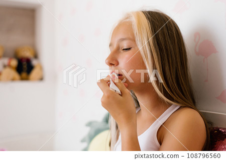 Caucasian teenage girl is sitting on the bed, looking in the mirror and painting her lips with balm 108796560