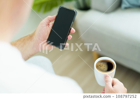 Caucasian white man wake up from the bed in morning and sitting beside the bed and holding an empty blank screen smartphone and enjoy drinking his morning coffee. Man checking message on smartphone. 108797252