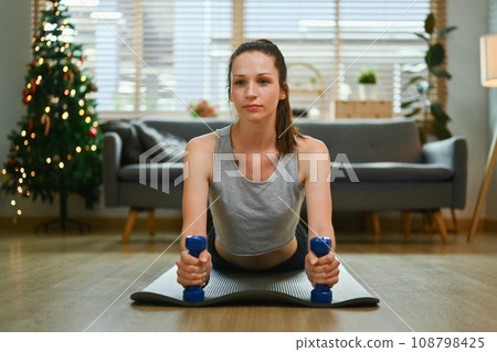 Concentrated young woman doing plank exercise on yoga mat during Christmas holidays. Healthy lifestyle concept Concentrated young woman doing plank exercise on yoga mat during Christmas holidays. Healthy lifestyle concept 108798425