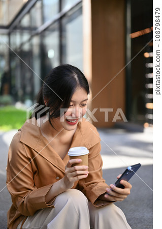 Young businesswoman with take away coffee cup in hand using mobile phone near office building Young businesswoman with take away coffee cup in hand using mobile phone near office building 108798479