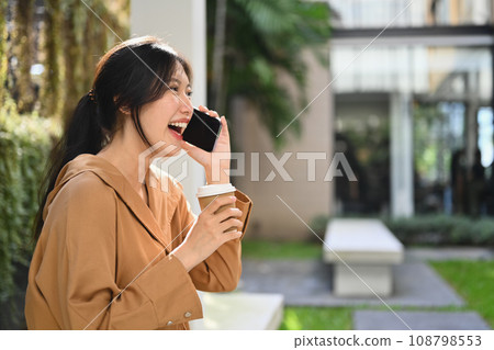 Businesswoman laughing and talking on mobile phone while sitting outside modern office building Businesswoman laughing and talking on mobile phone while sitting outside modern office building 108798553