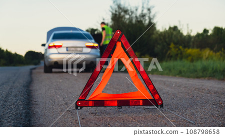 Broken silver luxury car emergency accident. Man driver installing red triangle stop sign on road. Sport automobile turned on blinkers technical problems on the road. Safety procedure vehicle broken 108798658