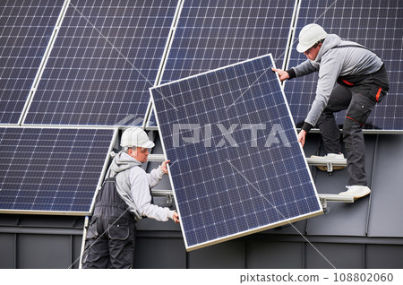 Builders building solar panel system on roof of house. Men workers in helmets carrying photovoltaic solar module outdoors. Concept of alternative and renewable energy. Builders building solar panel system on roof of house. Men workers in helmets carrying photovoltaic solar module outdoors. Concept of alternative and renewable energy. 108802060