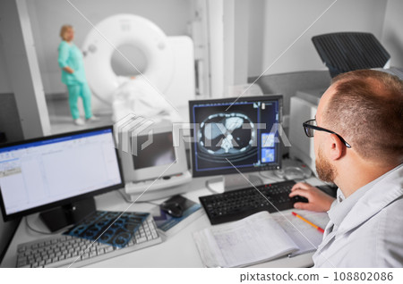 Medical computed tomography or MRI scanner. Back view of male doctor sitting at computer, nurse making MRI on blurred background. Specialists working in clinic. Concept of modern diagnostics. Medical computed tomography or MRI scanner. Back view of male doctor sitting at computer, nurse making MRI on blurred background. Specialists working in clinic. Concept of modern diagnostics. 108802086