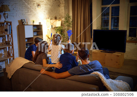 Back view. Family, parents and children sitting on couch in evening at home and watching football match translation. Cheering up team 108802506