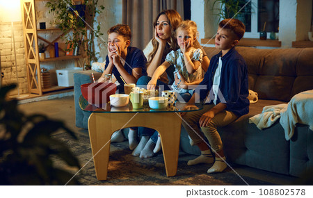 Young woman, mother spending time with her children, boys and little girl, family sitting on couch at home in evening and watching tv 108802578