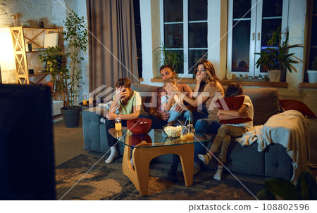 Family watching tv at home in the evening. Parents with children sitting on couch, watching horror movie, covering eyes in fear 108802596