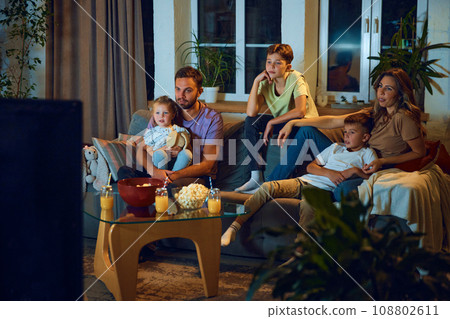Boys and girl, sitting on couch with parents, other and father in living room, watching TV, eating snacks in the evening Boys and girl, sitting on couch with parents, other and father in living room, watching TV, eating snacks in the evening 108802611
