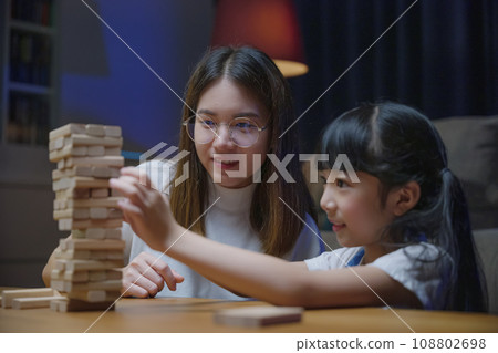 Asian young mother playing game in wood block with little daughter in home living room at night, Smiling woman help teach preschooler kid play build constructor tower of wooden blocks, family funny Asian young mother playing game in wood block with little daughter in home living room at night, Smiling woman help teach preschooler kid play build constructor tower of wooden blocks, family funny 108802698