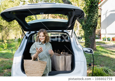 Woman with shopping bag next to a charging electric car Woman with shopping bag next to a charging electric car 108802759
