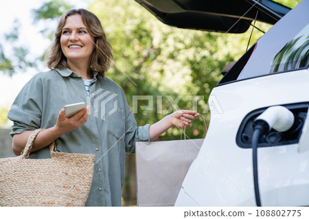 Woman with shopping bag next to a charging electric car 108802775