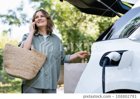 Woman with shopping bag next to a charging electric car 108802781