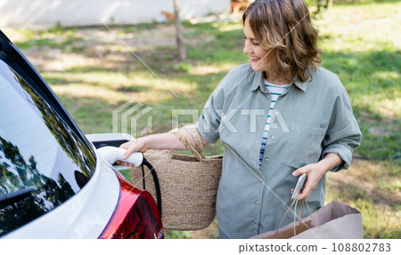 Woman with shopping bag next to a charging electric car 108802783