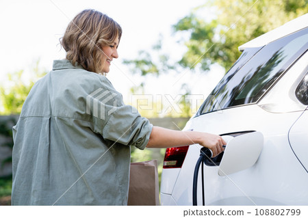 Woman with shopping bag next to a charging electric car 108802799